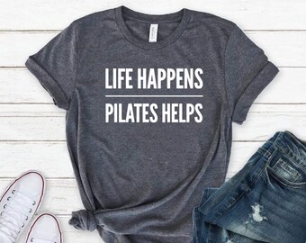 May include: A dark heather gray short-sleeve t-shirt featuring the white text "LIFE HAPPENS" above a horizontal line and the text "PILATES HELPS" below it. The shirt is displayed alongside a pair of white canvas sneakers and distressed blue denim jeans.