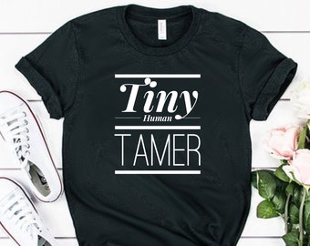 May include: A black short-sleeved t-shirt featuring white text that reads Tiny Human TAMER, with the word Tiny in a serif font and TAMER in a sans-serif font, separated by a horizontal line and the word Human in a smaller serif font.