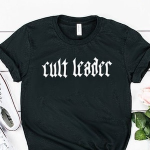 May include: Black t-shirt with white text that reads "cult leader" in a gothic font. The shirt is laid flat on a white wooden surface with a pair of white trainers and a bouquet of pink and white roses.