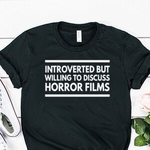 Może przedstawiać: Czarny t-shirt z kr&oacute;tkim rękawem z białym napisem "INTROVERTED BUT WILLING TO DISCUSS HORROR FILMS" na klatce piersiowej. Koszulka ma okrągły dekolt i jest pokazana obok białych trampek oraz bukietu r&oacute;żowych r&oacute;ż.