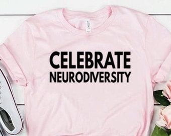 May include: A light pink short-sleeved t-shirt featuring the black text CELEBRATE NEURODIVERSITY printed on the front. The shirt has a crew neckline and is displayed with white canvas sneakers and a bouquet of pink and white artificial roses.