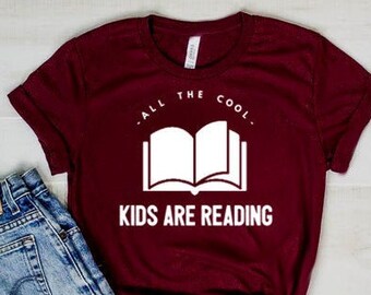 May include: A maroon t-shirt with the text "ALL THE COOL KIDS ARE READING" in white, along with a white graphic of an open book. The shirt is paired with blue denim jeans.