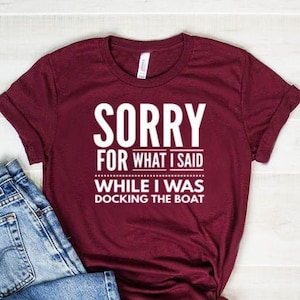 May include: A maroon t-shirt with white text that reads "Sorry for what I said while I was docking the boat". The shirt is laying on a light wood surface with a pair of blue jeans folded next to it.