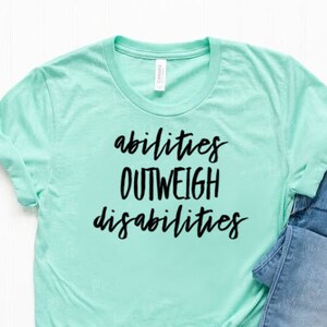 May include: A turquoise blue t-shirt with the text "abilities outweigh disabilities" in black lettering. The shirt is paired with blue jeans and white sneakers.