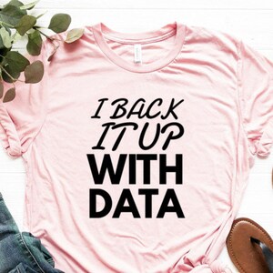 May include: A light pink short-sleeved t-shirt featuring the black text I BACK IT UP WITH DATA printed on the front. The shirt is displayed alongside a pair of blue denim jeans with distressed details and a pair of black strappy sandals.