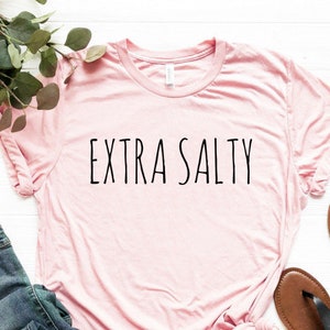 May include: A pink t-shirt with the text "EXTRA SALTY" printed in black on the front. The shirt is laying on a white surface with a pair of blue jeans and brown sandals.