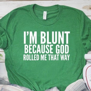 May include: A green t-shirt with the text "I'M BLUNT BECAUSE GOD ROLLED ME THAT WAY" in white. The shirt is paired with a plaid shirt and white canvas sneakers with red and blue accents.