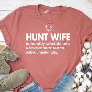 May include: A red t-shirt with the text "HUNT WIFE (n.) Incredibly patient; Married to a dedicated hunter; Seasonal widow; Ultimate trophy." in white text.