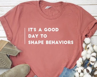 May include: A rust-coloured t-shirt with the text "It's a good day to shape behaviours" in white.