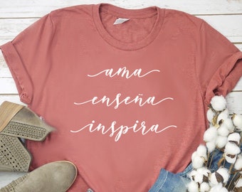 Puede incluir: Una camiseta rosa polvorienta con las palabras "ama enseña inspira" en escritura blanca. La camiseta se combina con vaqueros azules, botines grises y una ramita de algodón. Los artículos están dispuestos sobre una superficie de madera blanca.