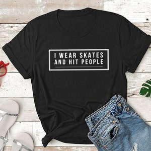 K&ouml;nnte beinhalten: Schwarzes T-Shirt mit wei&szlig;em Text, der "I wear skates and hit people" lautet. Das Shirt liegt auf einer wei&szlig;en Holzoberfl&auml;che mit einem Paar silbernen Sandalen, einer roten Sonnenbrille und einer blauen Jeans-Shorts.
