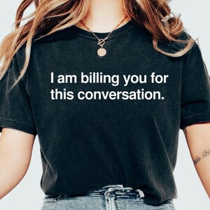 Może przedstawiać: Kobieta w czarnym t-shircie z napisem "Fakturuję ci tę rozmowę". Ma na sobie niebieskie dżinsy i białe buty sportowe.