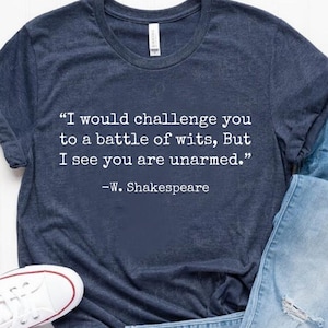Könnte beinhalten: Ein marineblaues T-Shirt mit einem weißen Zitat von William Shakespeare, das lautet: "Ich würde dich zu einem Kampf der Geister herausfordern, aber ich sehe, dass du unbewaffnet bist." Das Zitat ist mittig auf dem Shirt platziert.