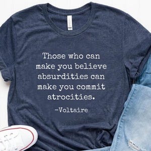 May include: Navy blue t-shirt with the quote "Those who can make you believe absurdities can make you commit atrocities. -Voltaire" in white text. The shirt is paired with light blue jeans and white Converse sneakers.