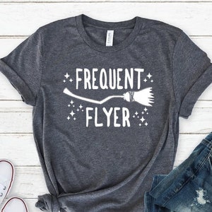 May include: A dark heather grey t-shirt with a white graphic of a broom and the text "Frequent Flyer".