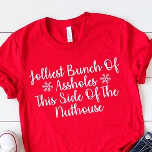 May include: A red t-shirt with the text "Jolliest Bunch Of Assholes This Side Of The Nuthouse" in white, paired with blue jeans and white sneakers. The shirt has rolled-up sleeves and a casual style.