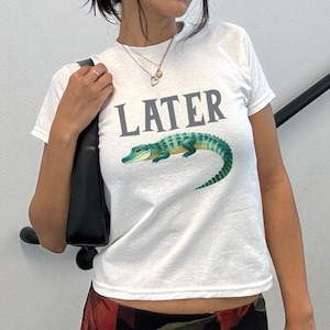 May include: White t-shirt with a green alligator graphic and the word "LATER" printed in gray. The model is wearing black sunglasses and a black floral skirt.