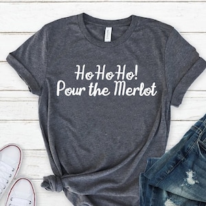 May include: A dark heather gray short-sleeve t-shirt featuring the white text "HoHoHo! Pour the Merlot" printed on the front. The shirt has a crew neckline and is displayed alongside a pair of blue denim jeans and white canvas sneakers.