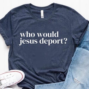 May include: A navy blue t-shirt with the text "who would jesus deport?" in white, paired with light blue denim jeans and white canvas sneakers.