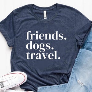 Puede incluir: Camiseta azul marino con las palabras "friends. dogs. travel." en blanco, combinada con vaqueros azul claro y zapatillas blancas. El conjunto informal está sobre una superficie blanca.