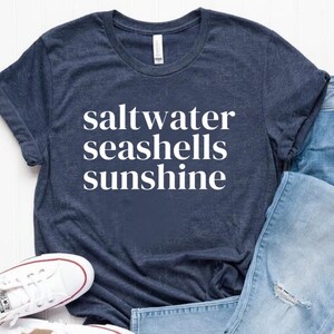 Op de afbeelding: Een marineblauw t-shirt met witte tekst die "saltwater seashells sunshine" zegt. Het shirt wordt gecombineerd met een blauwe jeans en witte sneakers.