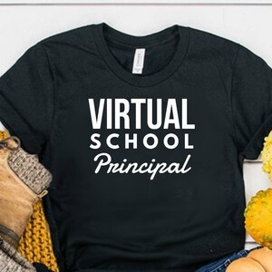 May include: A black short-sleeved t-shirt with the white text VIRTUAL SCHOOL Principal printed on the front. The shirt is displayed alongside a pair of brown lace-up boots, a mustard yellow knit scarf, blue denim jeans, and several small gourds.