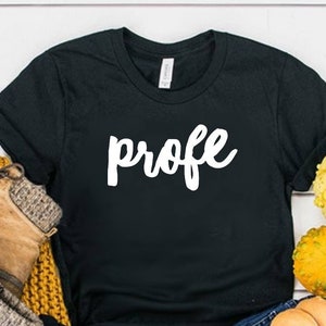 May include: Black t-shirt with the word "profe" written in white cursive lettering. The shirt is laid out on a white wooden surface with a pair of brown boots, blue jeans, and gourds.