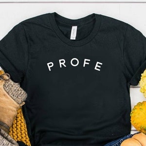 May include: A black short-sleeved t-shirt with the word PROFE printed in white capital letters across the chest, paired with blue denim jeans, tan lace-up ankle boots with grey knit cuffs, a mustard yellow knit scarf, and assorted small gourds.