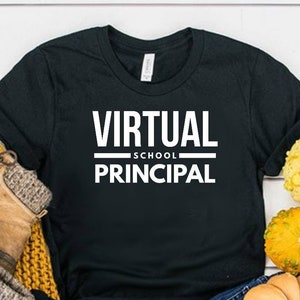 May include: A black short-sleeved t-shirt with the white text VIRTUAL SCHOOL PRINCIPAL printed on the front. The shirt is displayed alongside a pair of brown lace-up boots, a mustard yellow knit scarf, blue denim jeans, and several small gourds.