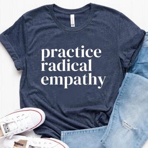 May include: A navy blue t-shirt with the text "practice radical empathy" printed in white on the front. The shirt is paired with blue jeans and white sneakers.