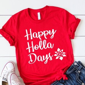 May include: A bright red short-sleeve t-shirt featuring the white text Happy Holla Days and a white poinsettia graphic. The shirt is paired with dark blue denim jeans and white canvas sneakers with white laces and rubber soles.