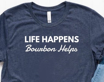 May include: A navy blue t-shirt with the text "Life Happens Bourbon Helps" in white lettering. The shirt is laid flat on a white surface with a pair of blue jeans and white sneakers.