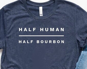 Könnte beinhalten: Ein marineblaues T-Shirt mit weißem Text, der "HALF HUMAN HALF BOURBON" lautet. Das Shirt liegt flach auf einer weißen Oberfläche mit einer blauen Jeans und weißen Turnschuhen.
