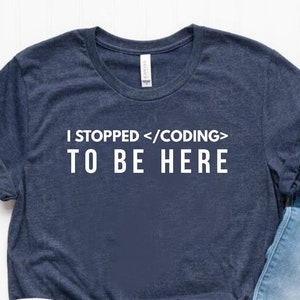 May include: A navy blue short-sleeved t-shirt with the white text I STOPPED </CODING> TO BE HERE, paired with light blue distressed denim jeans and a pair of white low-top canvas sneakers with red and blue trim along the rubber soles.