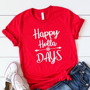 May include: A bright red short-sleeved t-shirt featuring the white text Happy Holla DAYS with a snowflake icon centered on an arrow graphic. The shirt is paired with dark blue denim jeans and a pair of white low-top canvas sneakers.