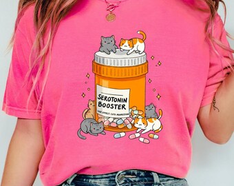 May include: Pink t-shirt with a graphic of a cartoon pill bottle labeled "Serotonin Booster" with cute cartoon cats surrounding it. The cats are various colors, including gray, orange, and white.