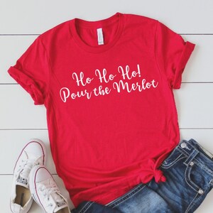 May include: A bright red short-sleeved t-shirt featuring the white cursive text Ho Ho Ho! Pour the Merlot, paired with dark blue denim jeans and white canvas sneakers with red and blue trim, arranged on a white wooden surface.