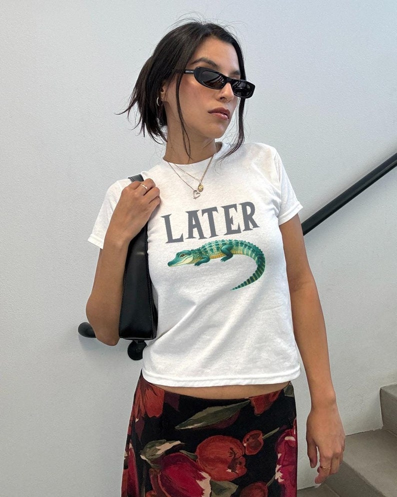 May include: White t-shirt with a green alligator graphic and the word "LATER" printed in gray. The model is wearing black sunglasses and a black floral skirt.