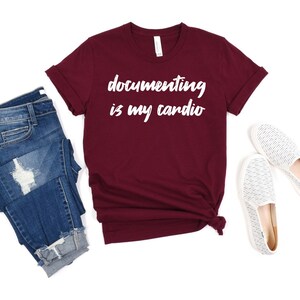 May include: A maroon short-sleeved t-shirt with the white text "documenting is my cardio" printed on the front, paired with blue denim jeans featuring distressed details and a pair of white slip-on shoes with a subtle grey patterned texture.