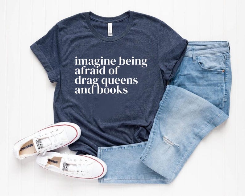 May include: A navy blue t-shirt with the text "imagine being afraid of drag queens and books" printed in white.