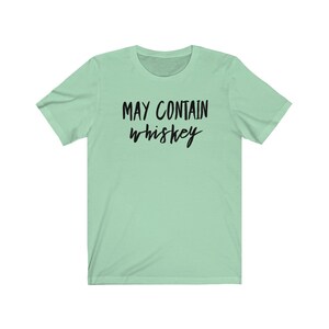 May include: A light green t-shirt with the text "MAY CONTAIN whiskey" in black script. The shirt has short sleeves and a crew neck. The text is centered on the front of the shirt.
