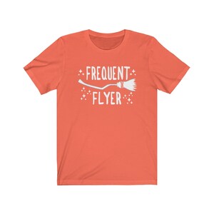 May include: A coral orange t-shirt with a white graphic of a broom and the text "Frequent Flyer".