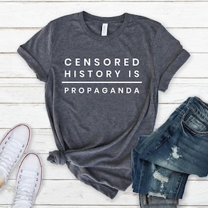 以下が含まれることがあります： 白い文字で「CENSORED HISTORY IS PROPAGANDA」と書かれた濃いグレーのTシャツ。白い木製の表面に白いスニーカーと青いジーンズと一緒に置かれています。