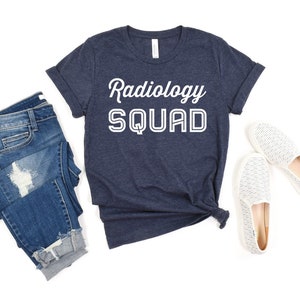 May include: A navy blue t-shirt with the text "Radiology Squad" in white lettering. The shirt is folded and laid flat on a white background with a pair of white jeans and white shoes.