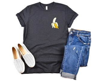 May include: A dark charcoal gray short-sleeve t-shirt featuring a small graphic of a partially peeled yellow banana on the left chest, paired with blue denim jeans and a pair of white slip-on shoes with a fine black horizontal line pattern.