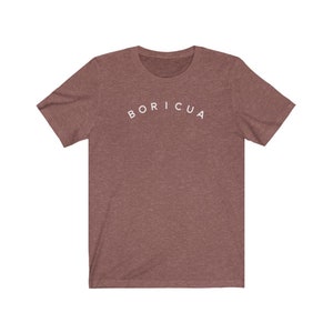Puede incluir: Una camiseta marr&oacute;n jaspeada de manga corta con la palabra "BORICUA" arqueada en letras blancas en el pecho. La camiseta est&aacute; hecha de un material suave y tiene un cuello redondo cl&aacute;sico.
