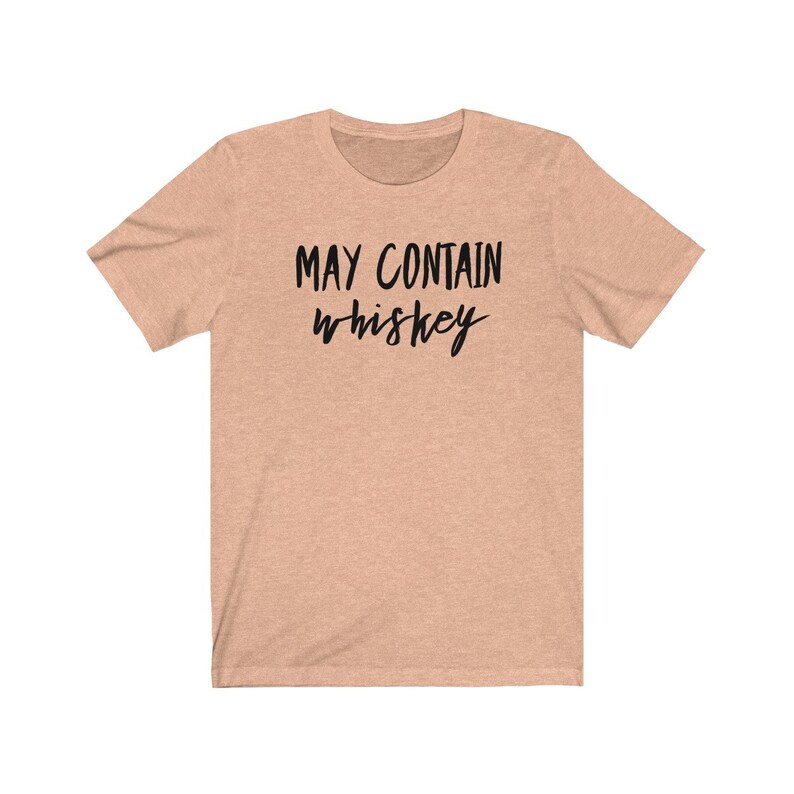 May include: A light peach-colored t-shirt with the text "MAY CONTAIN whiskey" in black, handwritten-style font. The shirt has short sleeves and a crew neck.