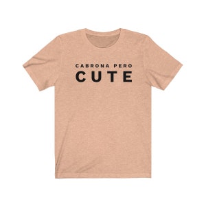 May include: A light peach colored t-shirt with black text that reads "Cabrona pero cute".