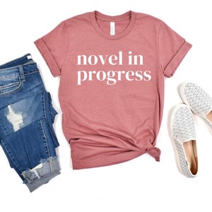 May include: A dusty rose short-sleeve t-shirt with the white text "novel in progress" printed on the front, paired with blue denim jeans featuring a distressed knee and a pair of white slip-on shoes with a fine black horizontal line pattern.