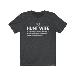 May include: Dark heather grey t-shirt with a white graphic that reads "HUNT WIFE (n.) Incredibly patient; Married to a dedicated hunter; Seasonal widow; Ultimate trophy."